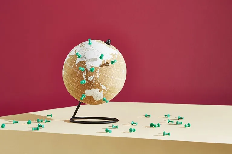 A photo of a world globe marked with pins on a desk in front of a red background