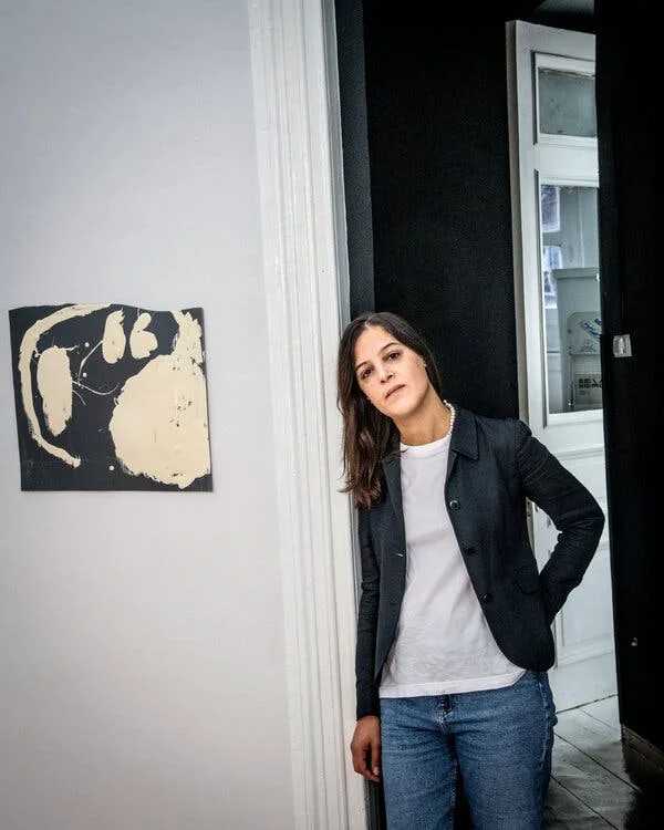 A woman, wearing bluejeans, a white T-shirt and a black jacket, leans on a doorway in an art gallery.