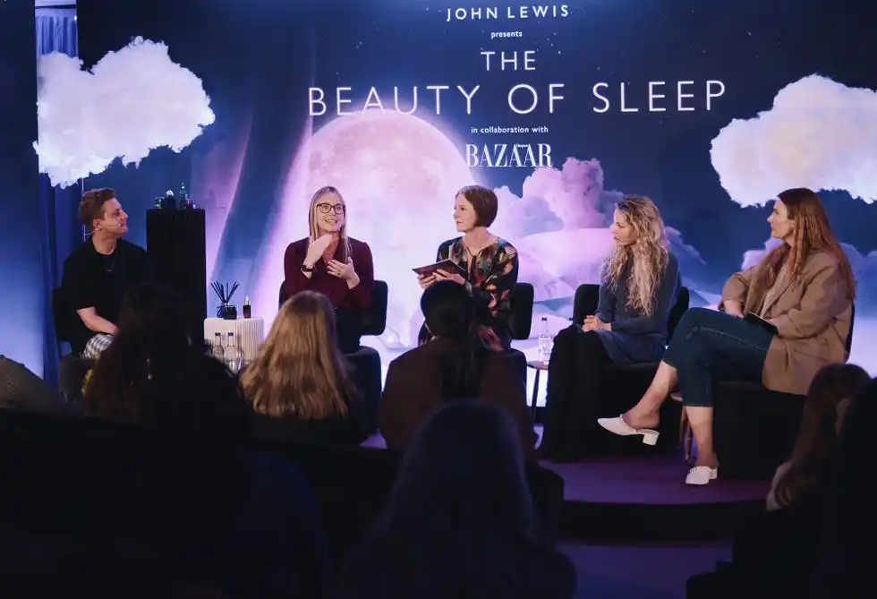 a panel on sleep wellness featuring multiple speakers seated in front of a large backdrop displaying the text 'the beauty of sleep' various decorative elements, such as clouds, complement the theme the speakers are engaged in conversation, with some holding notes or discussing topics pertinent to sleep the audience is partially visible, indicating an interactive event