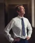 Andy Haldane stands in a dark corridor with the light focused on him; he has his hands on his hips and his face is turned almost in profile. He wears a bright white shirt and dark striped tie. 