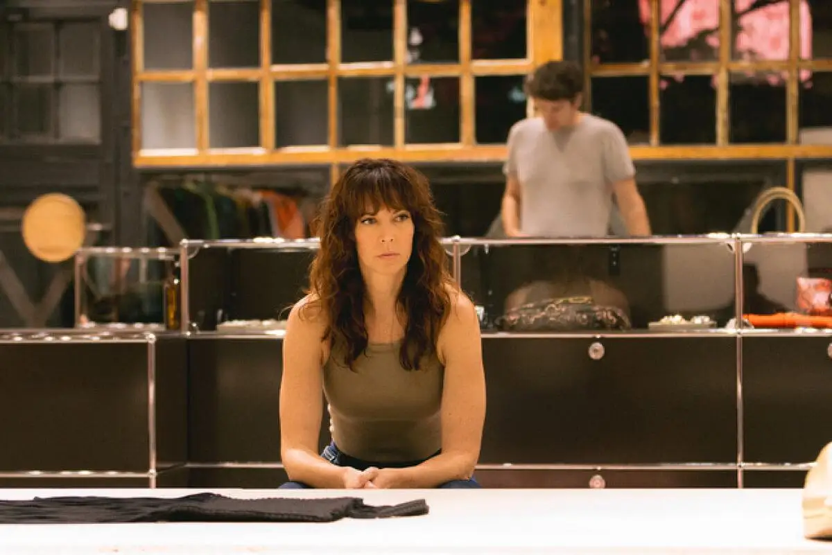 A serious-looking woman leans on a counter in a retail store. A man stands at a counter behind her in the distance.