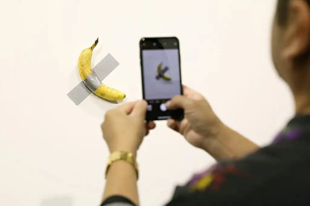 a person takes a picture of a banana taped to a wall with a smart phone