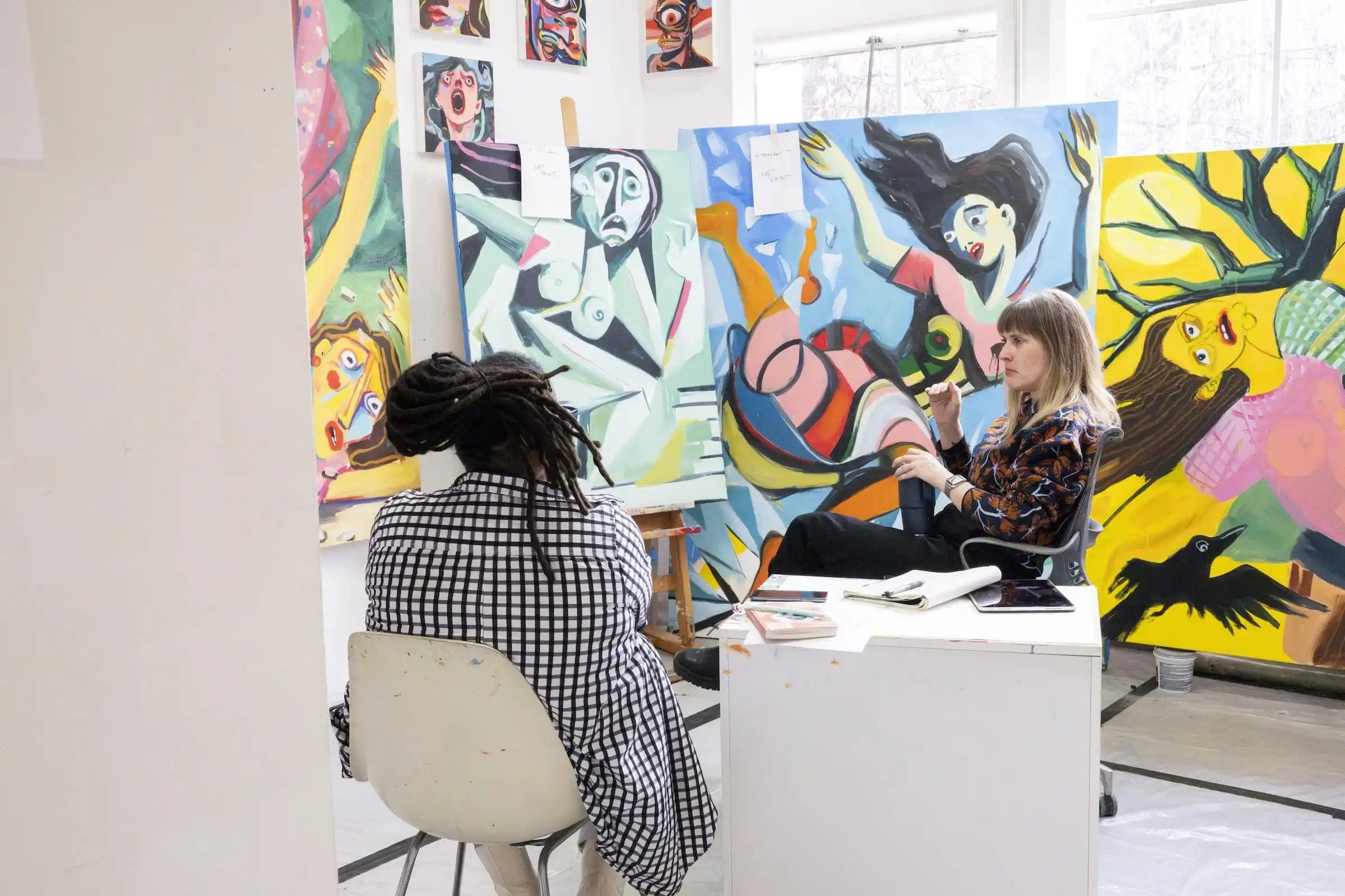 Two people sitting and conversing in front of bright, abstracted figurative paintings.