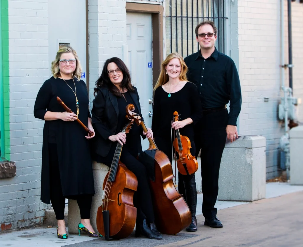 Four musicians in black outfits stand together, holding cellos and other instruments, representing Parish House Baroque.