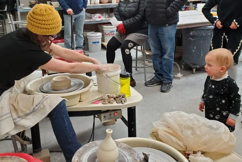 A person in a yellow hat and black T-shirt sits at a pottery wheel as a small child looks on.