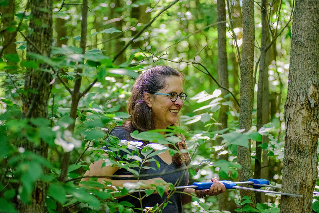 a woman with short curly hair is shown within a frame of bushes and trees in the woods.