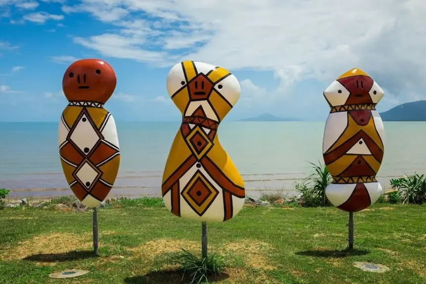 Three large body and head-shaped painted sculptures on metal poles, standing on grass with sea and islands behind.