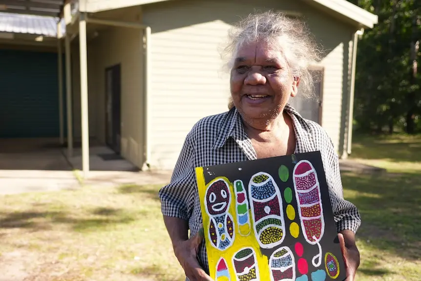 An elderly Aboriginal woman laughs while holding a painting.