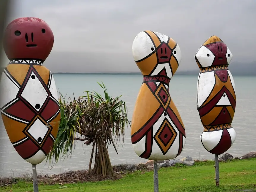 Three large body and head-shaped painted sculptures on metal poles, standing on grass with sea and islands behind.