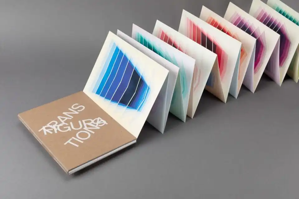 an accordion book with a kraft cover, which reads 'Transfigurations' in white, jumbled font, with pages of colorful geometric paintings