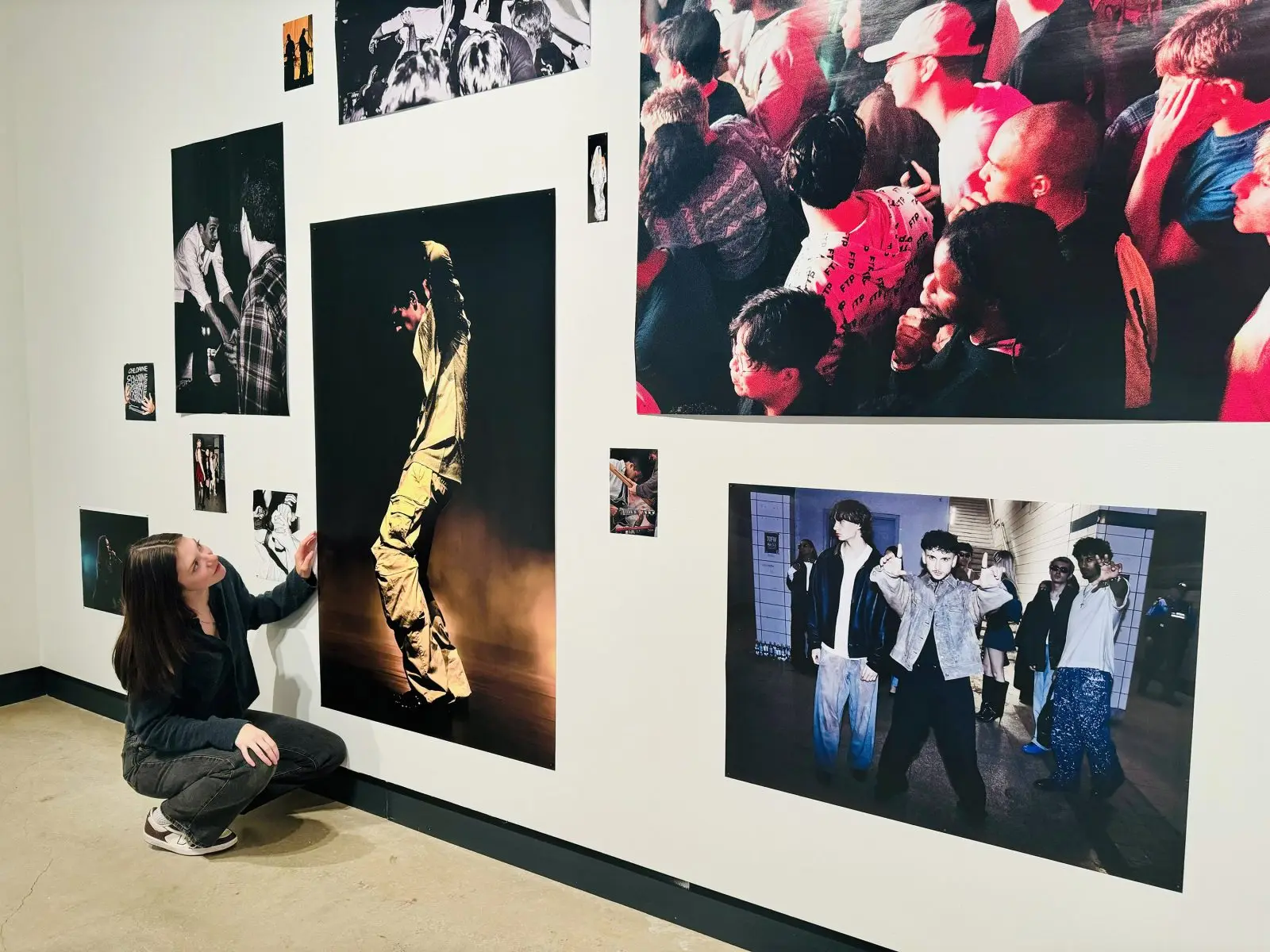 A woman crouches beside a wall of colourful photographs, looking up at a photo of a person at a music festival performing under stage light.