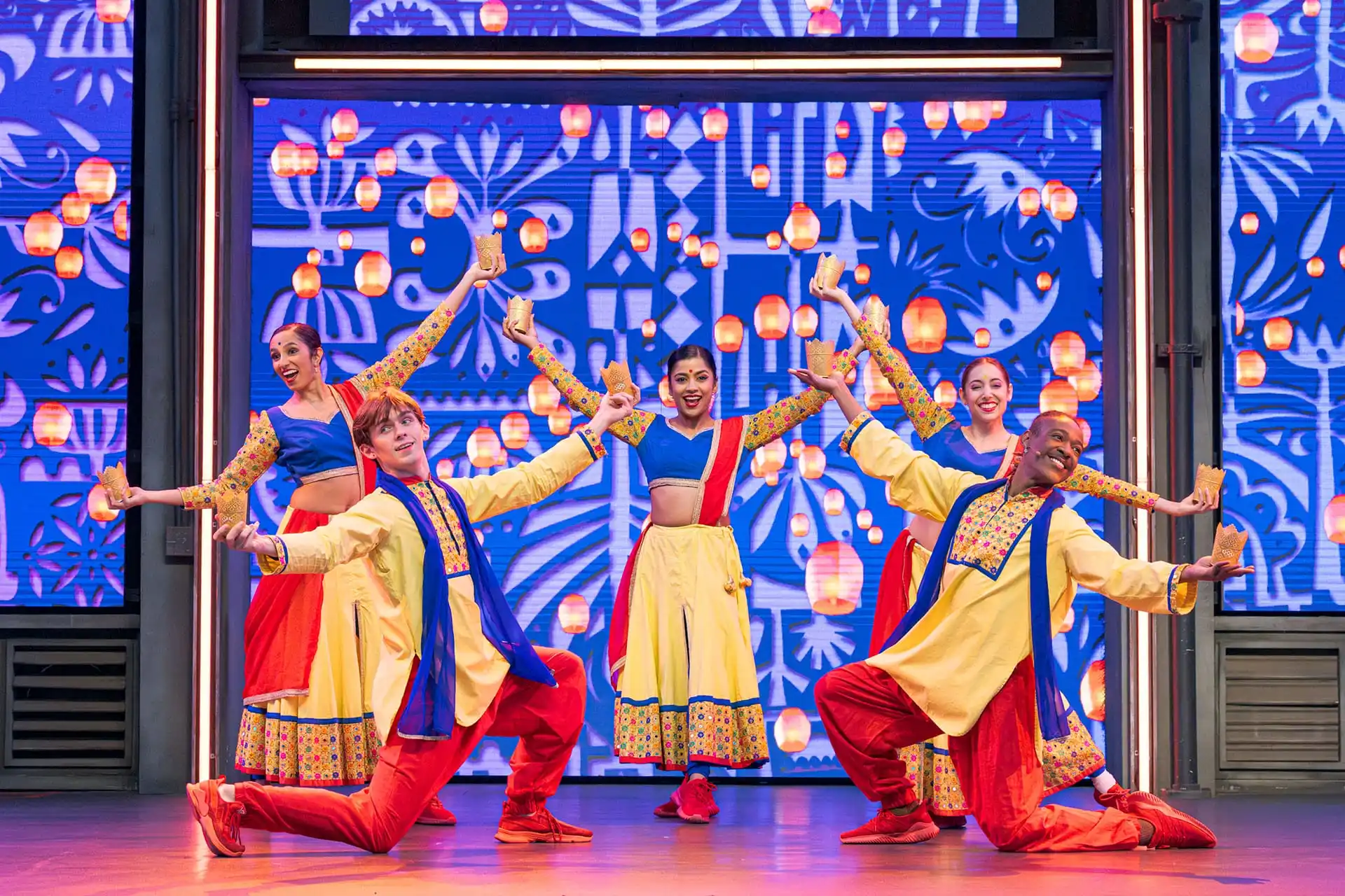 A group of dancers in colorful traditional attire performs on stage amid a vibrant patterned backdrop and hanging lanterns, offering enchanting holiday entertainment reminiscent of a Disneyland spectacle.