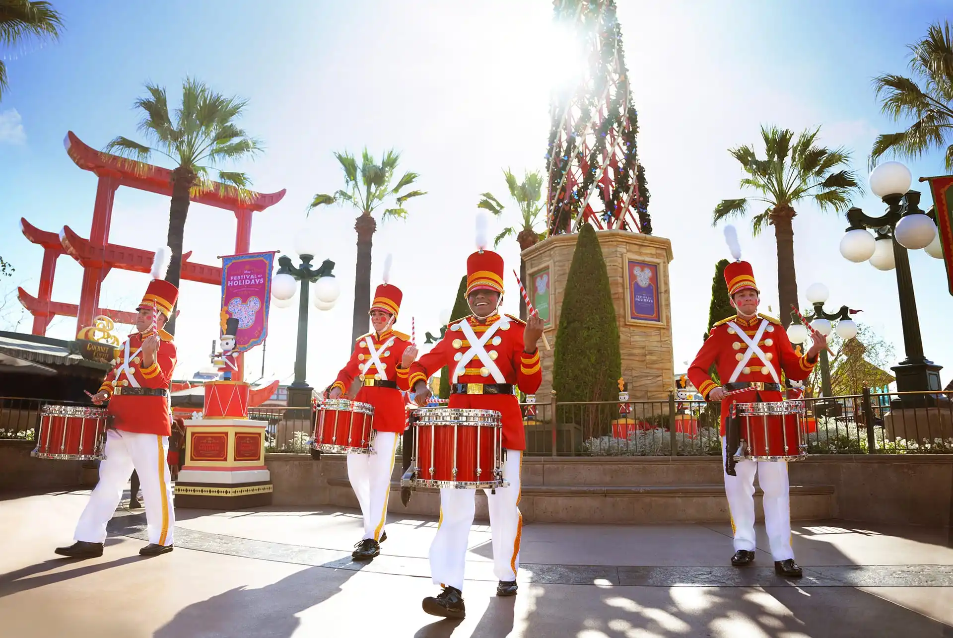 Four marching band drummers in red uniforms deliver holiday entertainment outdoors in a sunny amusement park setting, surrounded by palm trees and festive decorations.