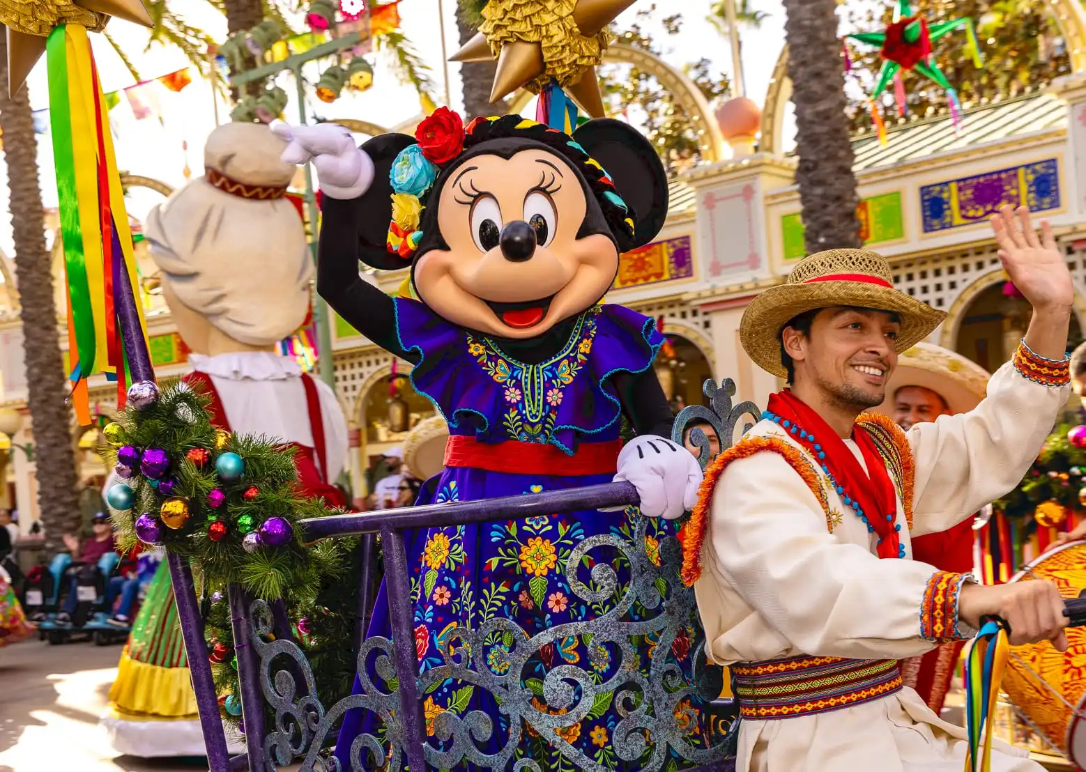 Minnie Mouse in a colorful dress waves from a Disneyland parade float, accompanied by performers in traditional attire during a festive holiday entertainment celebration adorned with vibrant decorations.