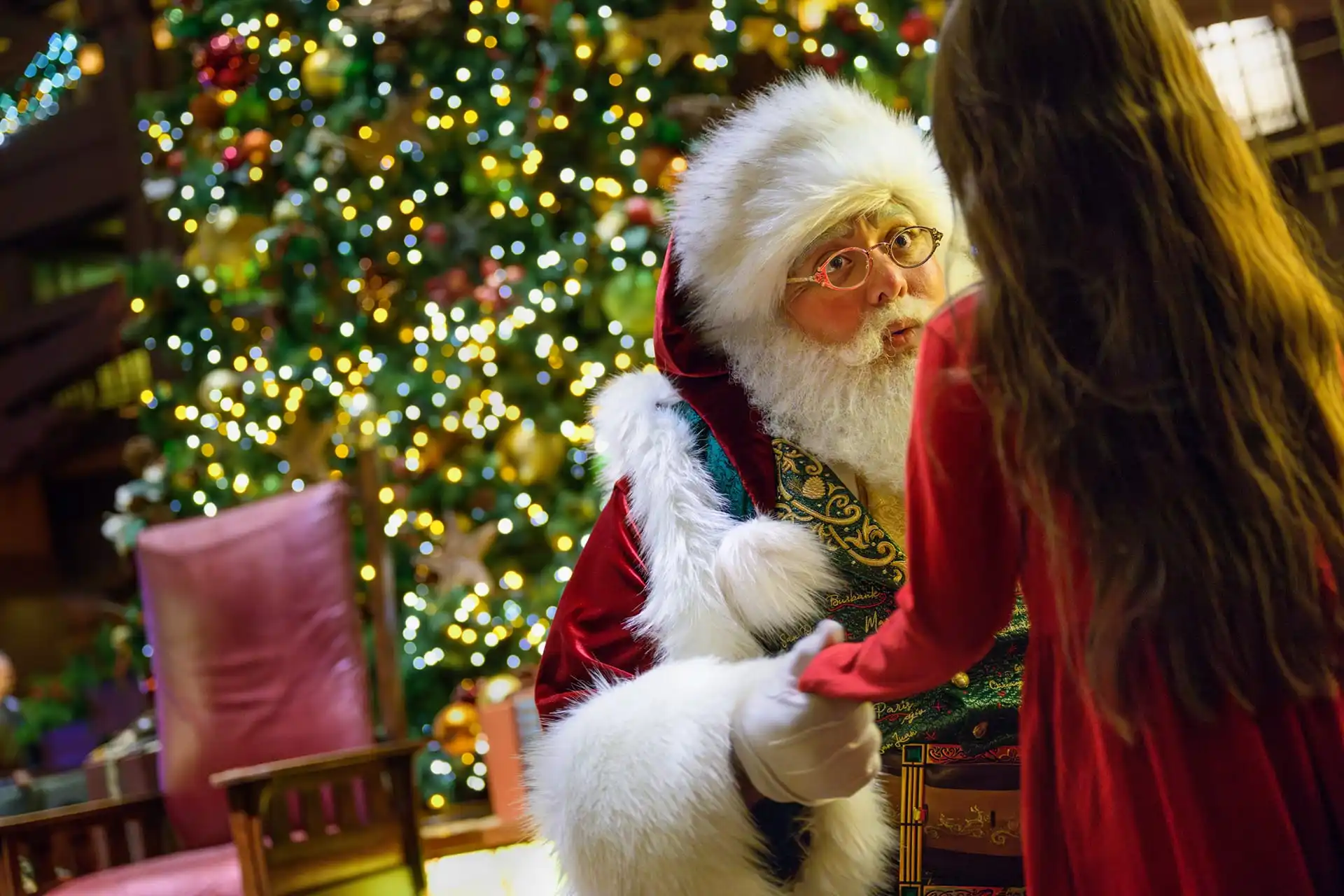 Santa Claus holds a child's hand in front of a large, decorated Christmas tree, creating a magical holiday entertainment scene reminiscent of Disneyland in a cozy setting.