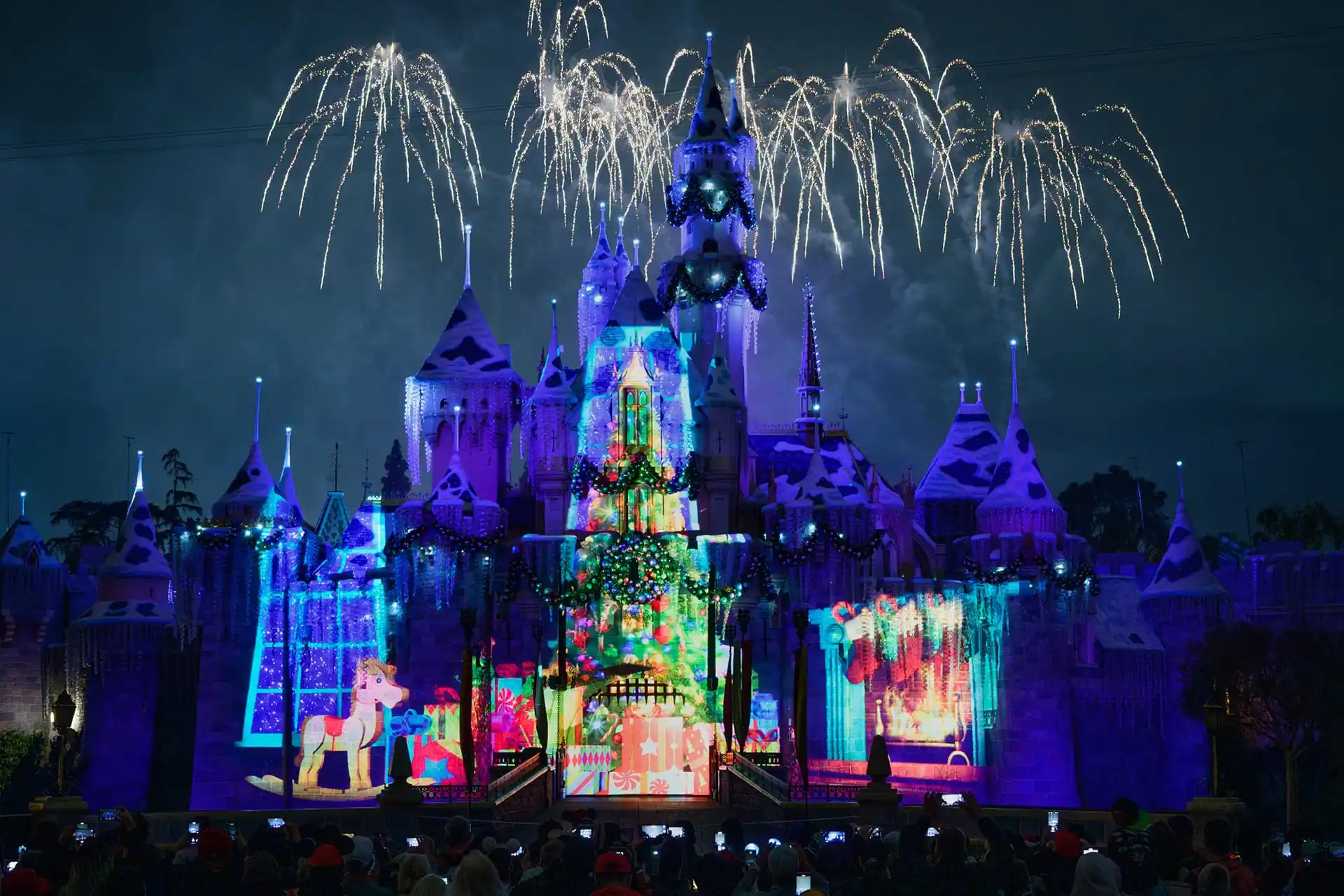 A brightly lit castle at Disneyland dazzles with holiday entertainment as Christmas projections dance across its walls and fireworks light up the night sky. A delighted crowd watches the spectacular display, captivated by the festive magic.