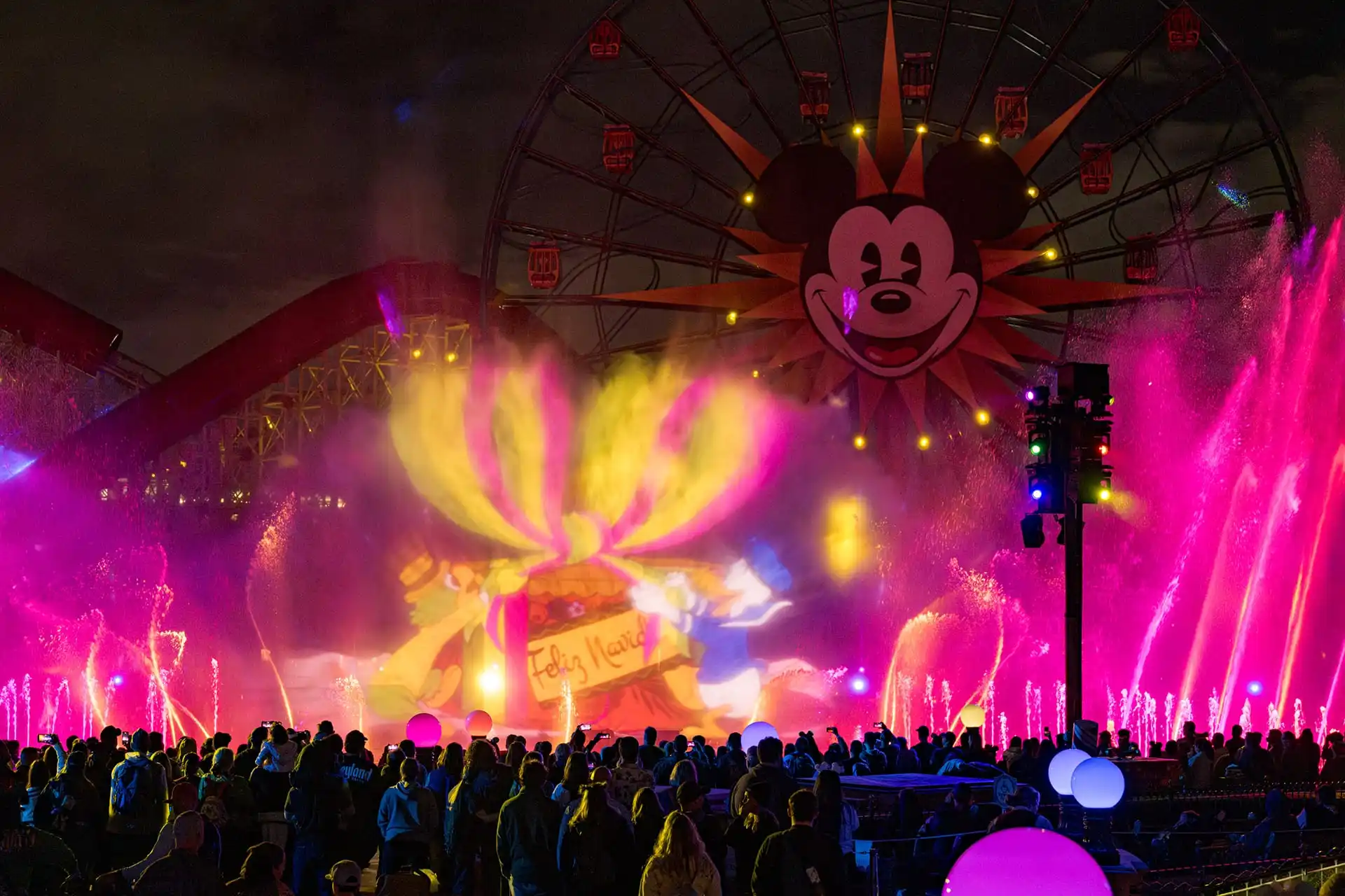 Crowd enjoying a nighttime light and water show at a theme park, where holiday entertainment comes alive with colorful projections and a Ferris wheel featuring a large cartoon face.