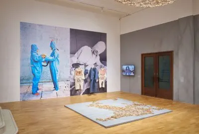 View of a museum exhibition showing various artworks including photographs printed onto walls of people wearing suits with the suits hanging on the walls. 