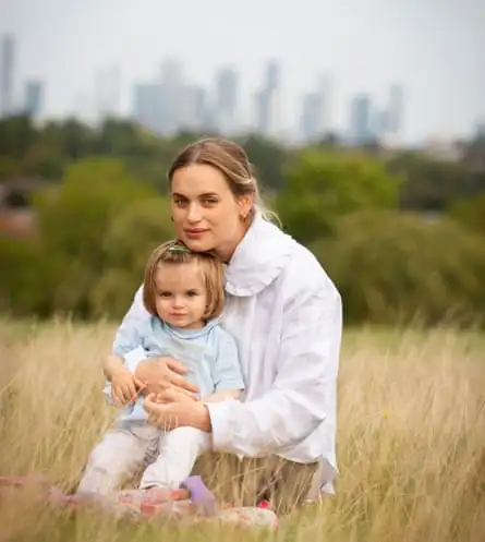 A woman with blond hair pulled back from her face and wearing a white blouse, holding a little girl with blond hair and wearing a pale blue top and striped leggings, sitting in long grass, with trees and tall buildings in the distance behind them