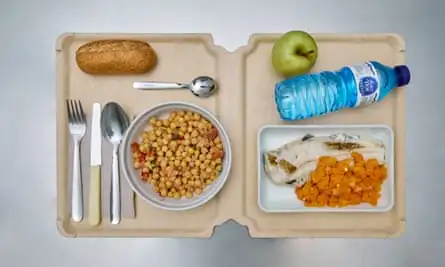 A tray with two sections, with cutlery, a bowl of chickpeas and a bread roll on one side, and a dish with a piece of fish and orange veg, a plastic bottle of water and an apple on the other