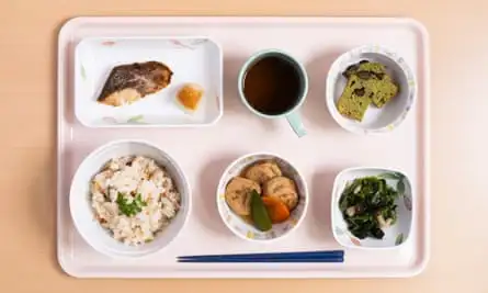 A plastic tray with chopsticks and bowls of rice, dumplings, vegetables, fish and cake, and a mug with a brown liquid in