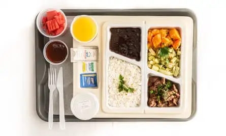 A plastic tray with a divided dish with rice and beans, vegetables, and some meat, fruit, water, juice and a dessert