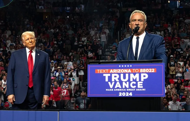 Old man with yellow hair, blue suite and red tie on the left and white hair guy in a blue suite speaking from a podium during a rally. Former presidential candidate Robert F. Kennedy Jr. (R) speaks as Republican presidential nominee, former U.S. President Donald Trump listens during a campaign rally at Desert Diamond Arena on August 23, 2024 in Glendale, Arizona.