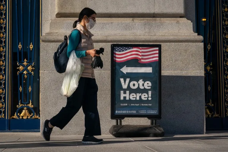 A person carrying a bag and wearing a face mask walks past a sign reading 'Vote Here'