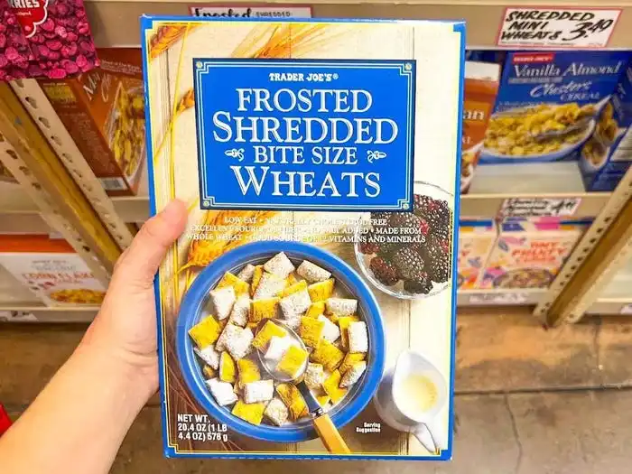 A hand holds a blue box of frosted bite-size wheats with an image of a bowl of cereal on the packaging