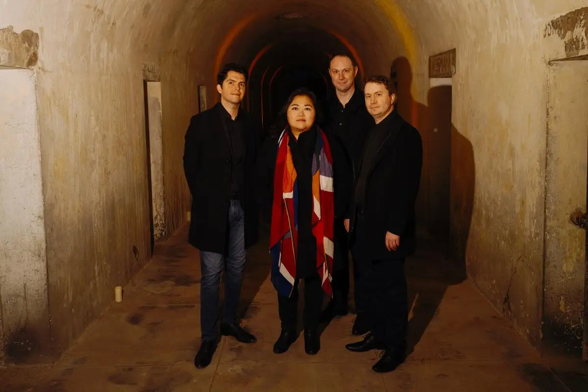 Three men and a woman, dressed mostly in black, stand under an arched ceiling