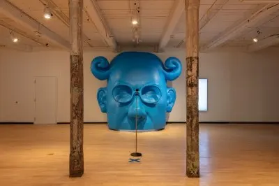 A large sculpture of part of Sir Cyril Radcliffe's face as a blue horned Dijnn. A microphone stand with a tan can stands in front of it. 