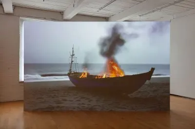 A screen showing a video of a boat run aground on the shore that is on fire. 