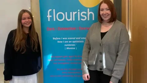 Evie, a girl supported by Flourish, stands on the left of a sign with the charity logo on. Flourish mentor Helen Laycock stands on the right of the sign.