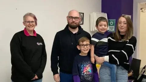 Four people stand in the Shine a Light charity building. On the left, founder Sam Schooler. Then two young boys called Archie and Leon, with their dad Niall and their mother Rhiannon, who is carrying Archie.