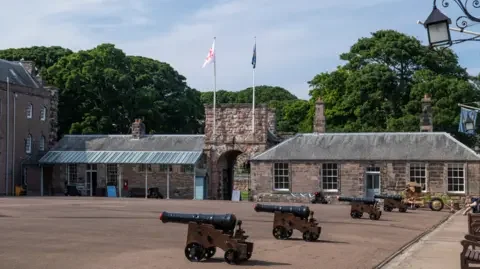 Maltings Trust Berwick parade ground, part of Berwick barracks with a row of field guns in the foreground and a low one story building behind them with an archway