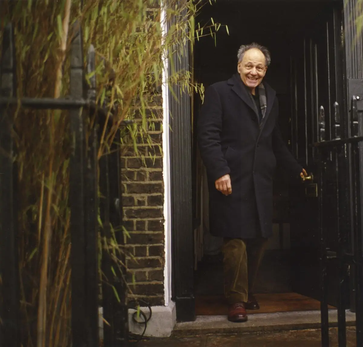 Artist Frank Auerbach in a dark coat and brown pants walking through an open door toward the camera.