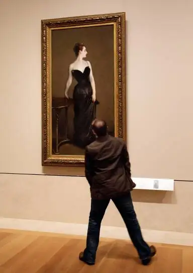 A man stands wide as he looks up at a painting of a woman.