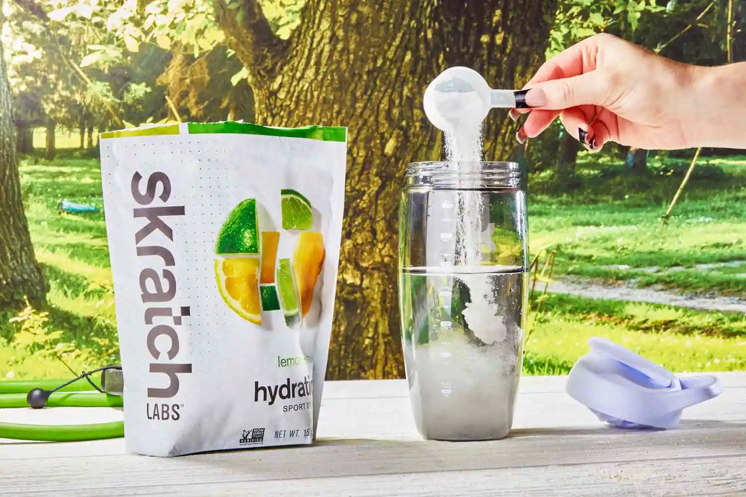 A hand pouring a scoop of Skratch Labs Sport Hydration Drink Mix into a glass bottle on a wooden table