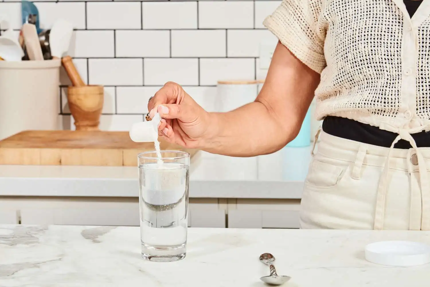 A hand using a scoop to add sports drink powder to a glass of water on a counter