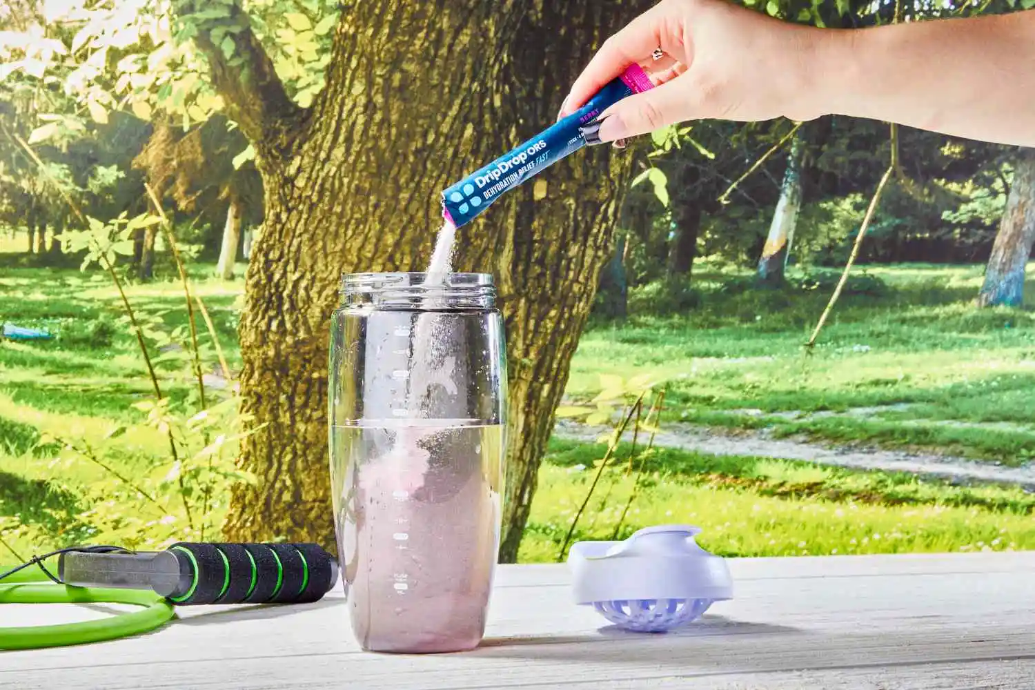 A hand adding a DripDrop Hydration Electrolyte Powder Packet into a glass on a wooden table outdoors