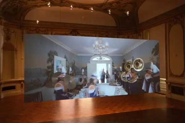 A hanging screen in an empty room, playing a video of a marching band performing around a dining room table.