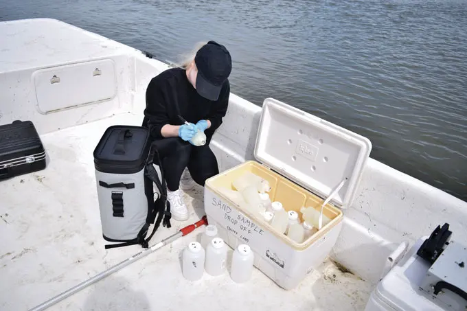 A woman in a baseball cap organizes containers of sand samples while on a boat