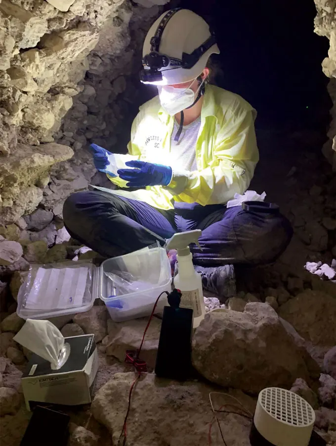 A woman sitting cross-legged in a cave surrounded by equipment and wearing gloves and a lit head lamp