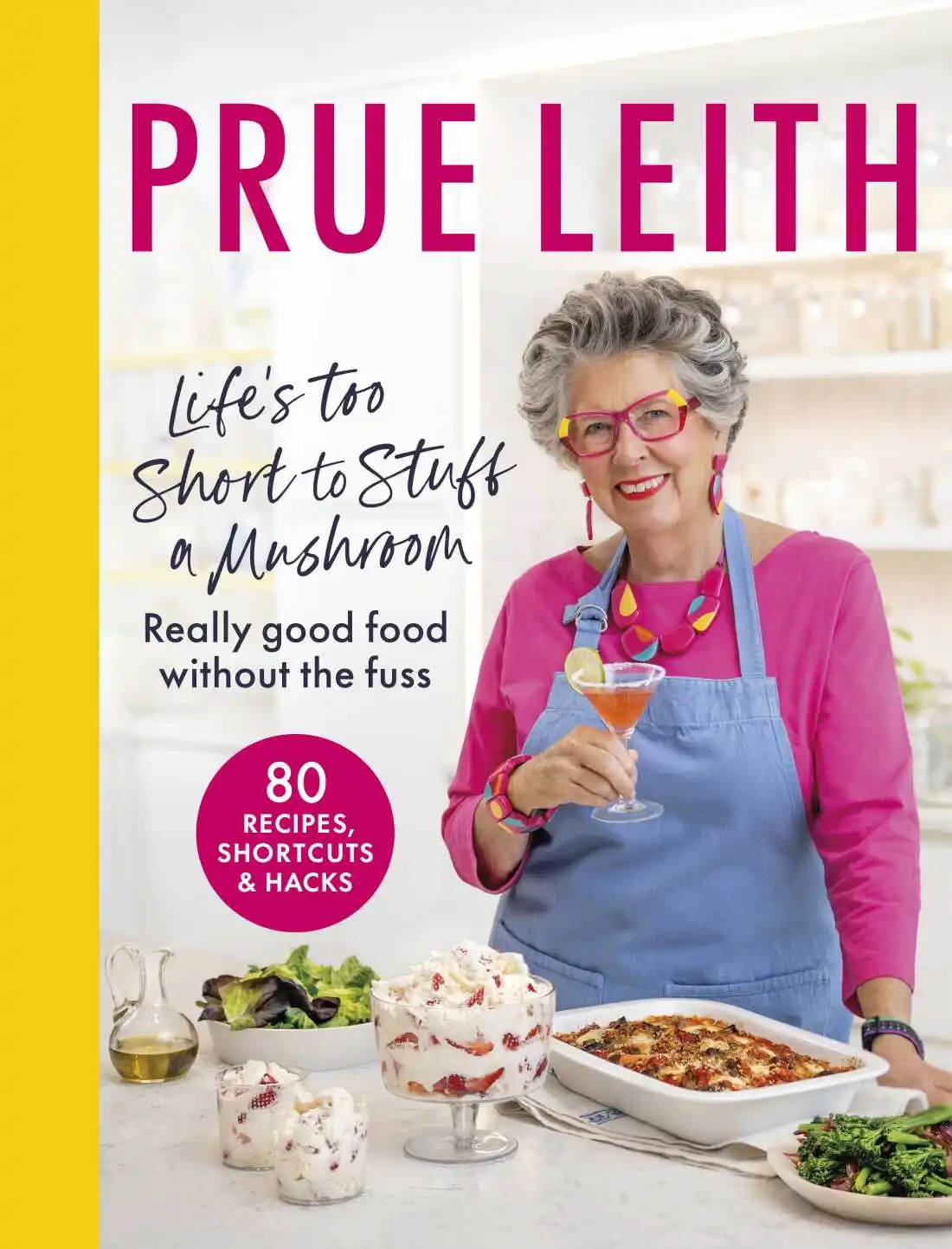 Photograph of the book cover of the cookbook, Life's Too Short To Stuff A Mushroom, by Prue Leith. The book cover depicts Prue in a kitchen, holding a drink and standing behind several prepared dishes on the counter in front of her. 