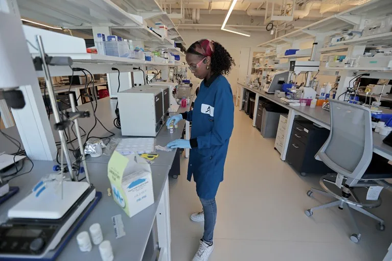 African American female wearing blue lab coat and blue gloves is standing at a lab work bench, focusing on some glass vials