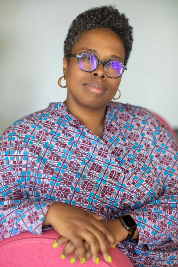 James wears a blue and pink patterned top. She sits in a pink chair, with her hands folded over the side. She has yellow fingernails. She wears glasses and has short hair and is looking at the camera.