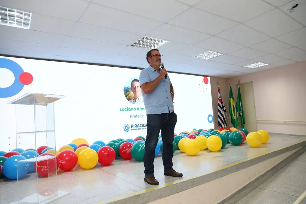 A imagem mostra Luciano Almeida, prefeito de Piracicaba, dando um discurso em um evento. Ele está usando uma camisa azul claro e calças jeans, e está segurando um microfone. Há uma tela grande atrás dele com seu nome e uma logo da prefeitura. O cenário inclui um palco pequeno com vários balões coloridos e bandeiras. 