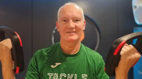 Paul Moseley/BBC Steve Stone - who has short white hair - wearing a green t-shirt with the word 'Tackle' on the front in white capital letters. He is holding a piece of workout equipment in each hand.