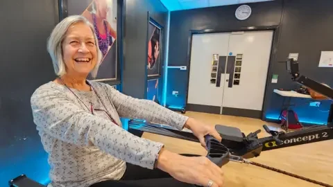 Paul Moseley/BBC Pam Jackson sitting on a rowing machine. She has silvery bobbed hair to just above her shoulders. She's smiling broadly whilst wearing a grey, patterned top