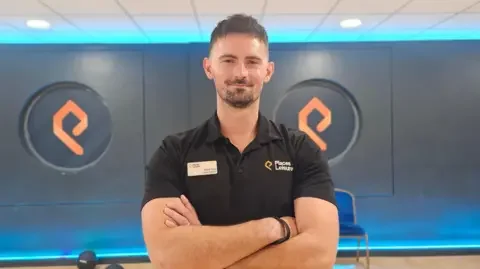 Paul Moseley/BBC David Lane smiling with his arms folded in the gym. He's wearing a black t-shirt and has short dark hair and a goatee beard